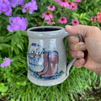 Rowe Pottery Hand thrown stoneware mug In a hand decorated Boots pattern being held up with flower background